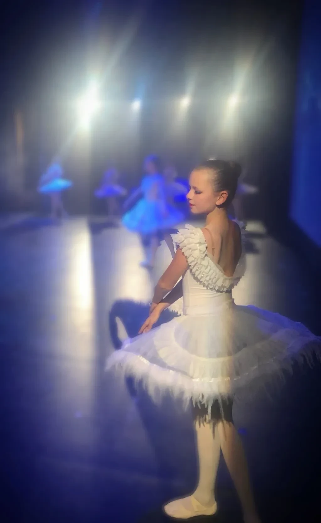 Young ballerina in white feathered tutu on blue-lit stage