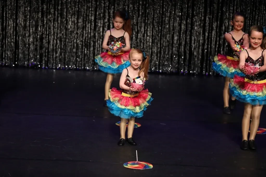 Young dancers in rainbow ruffled tutus performing on stage with lollipop props