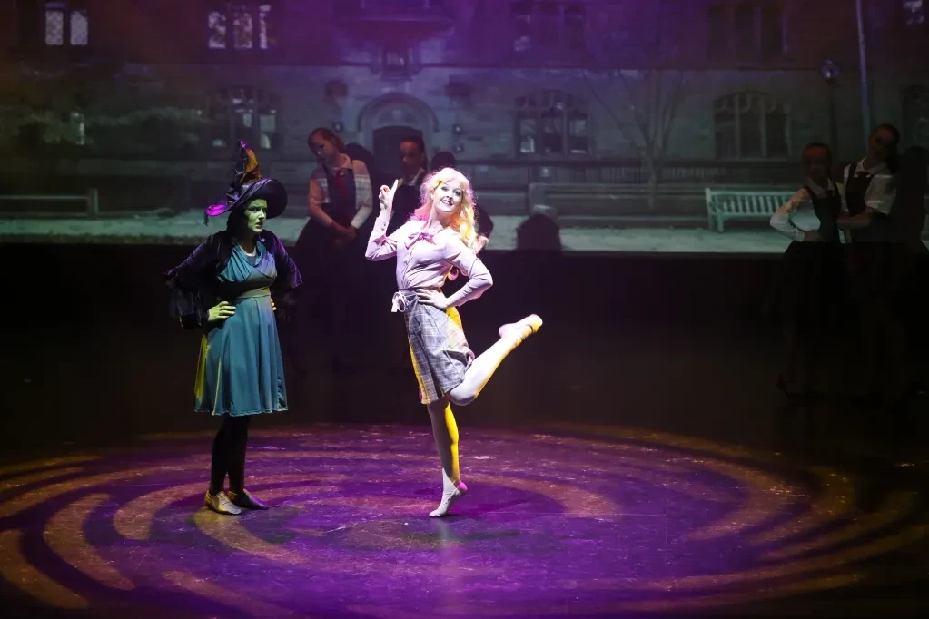 Wicked-themed scene with green witch and Glinda character kicking on purple-lit stage