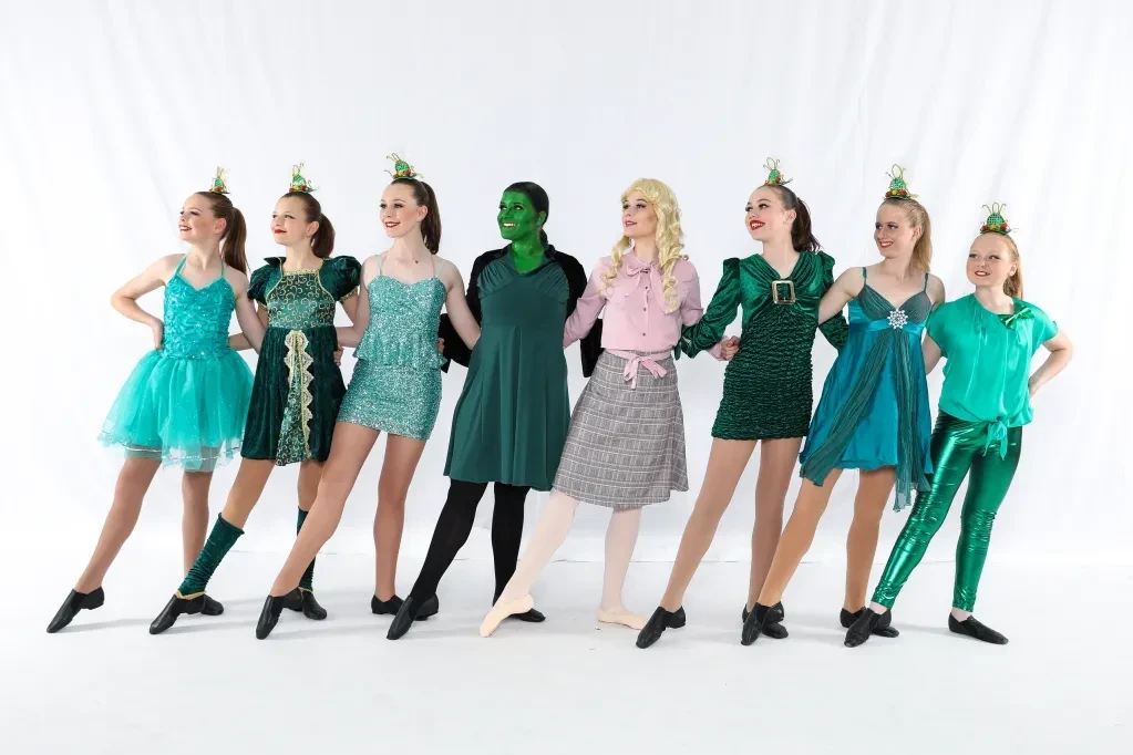 Eight dancers in green and teal Wicked costumes posed in a line on white background