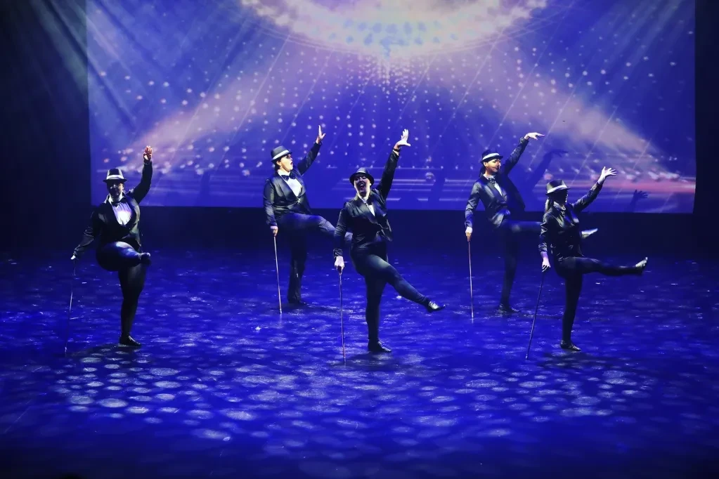 Five dancers in black tuxedos and top hats with canes on blue-lit stage