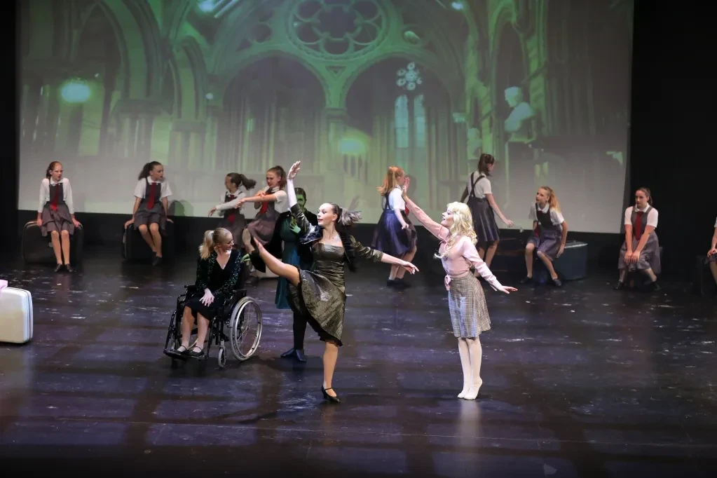 Full Wicked ensemble scene with wheelchair dancer, Elphaba and Glinda in cathedral backdrop