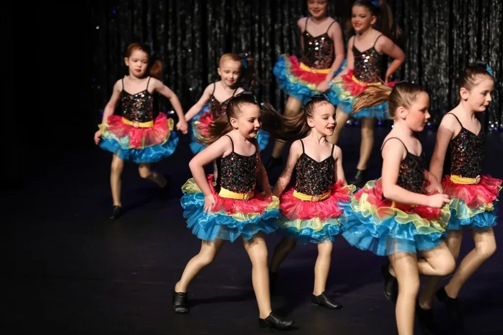 Group of young children in black sequin tops and rainbow tutus running across stage