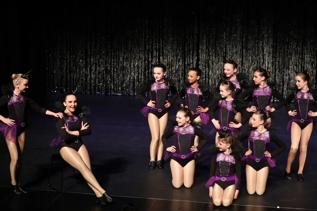Large group of dancers in black and purple jazz costumes posed on stage against silver curtain