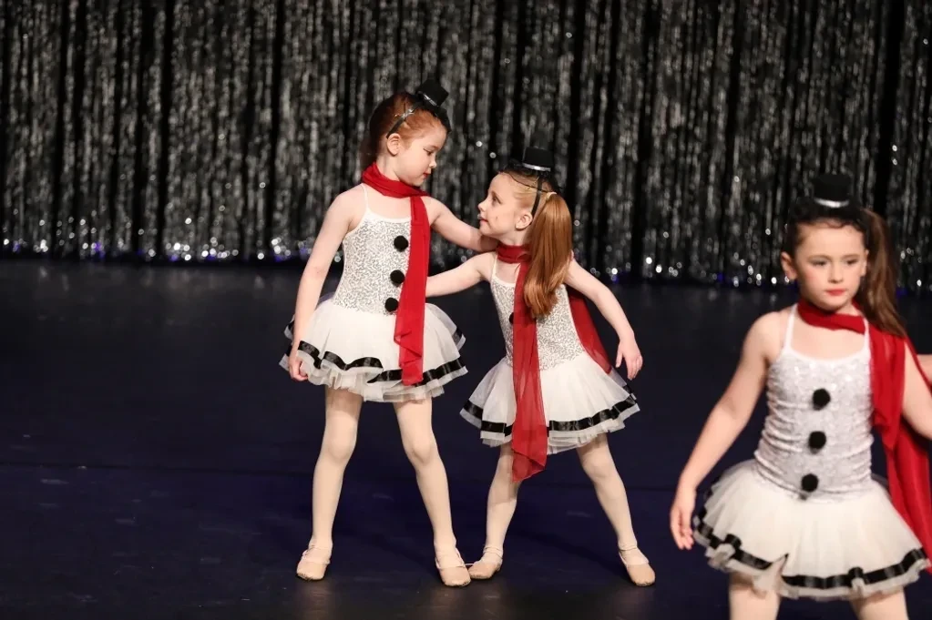 Three tiny dancers in white snowman costumes with red scarves and top hats on stage