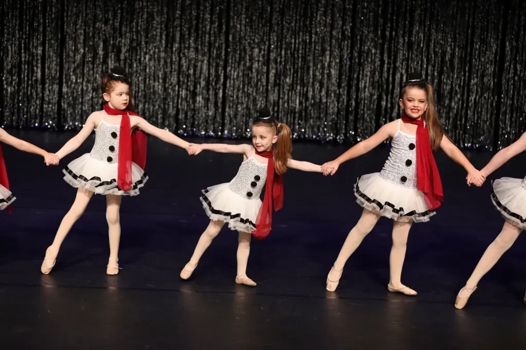 Young dancers in white snowman tutus with red scarves holding hands in a line on stage