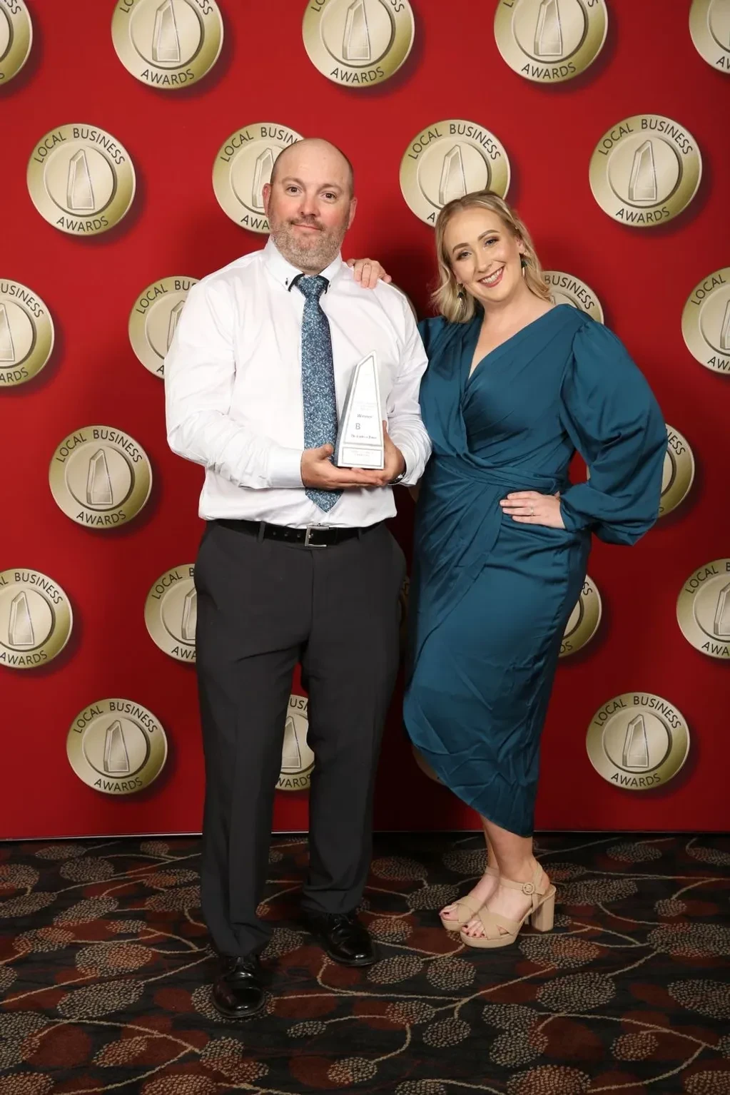 Two people posing with Local Business Awards trophy against red awards backdrop